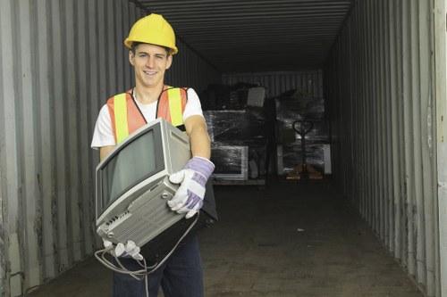 A young man wearing a yellow safety helmet, high-visibility vest, and white gloves during a rubbish removal task stands inside a metal storage container or industrial shed on a concrete surface. He is holding an old desktop computer with a CRT monitor and visible cords in both hands, with a slight smile on his face. The interior of the container is dimly lit, with a background showing stacked black plastic waste bags or tarps and a small manual pallet truck or hand truck positioned further inside. The scene suggests an environmental or waste management context, possibly part of a commercial rubbish disposal process in Fulham or nearby areas, highlighting the services offered by Commercial Waste Removal Fulham with a focus on thorough and professional rubbish clearance.