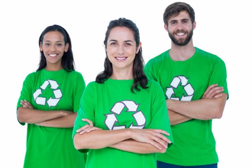 Three individuals wearing bright green T-shirts featuring a white recycling symbol stand outdoors on a paved surface, likely a driveway or street, with arms crossed and smiling at the camera. The person in the center, a woman with dark hair styled in loose waves, is positioned in the foreground. To her left, a woman with long dark hair and darker skin tone also appears in the background. To her right, a man with a beard and short dark hair is similarly positioned behind her. The vibrant T-shirts indicate their association with environmental or waste management initiatives, aligning with the context of rubbish removal services. The background is blurred but suggests an urban or suburban setting, possibly in Fulham, with natural lighting creating soft shadows and a clear, professional atmosphere. This image visually emphasizes teamwork and environmental responsibility, which are integral to commercial waste removal operations by companies like Commercial Waste Removal Fulham, serving the local community in the SW6 area.