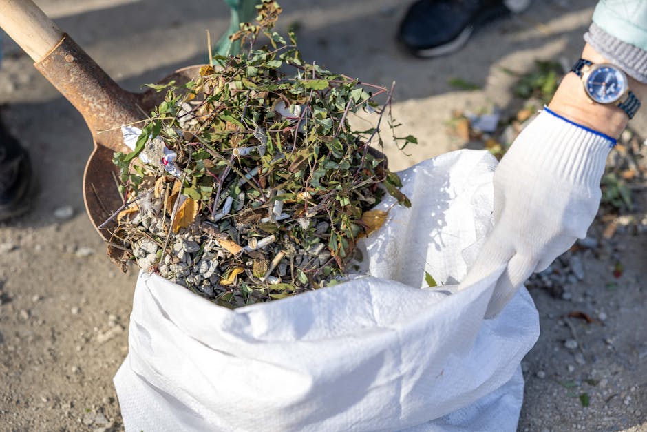 A person wearing a green long-sleeved shirt, a plaid jacket in shades of yellow, gray, and orange, and light-colored trousers is holding open a large black rubbish bag lined with a shiny black plastic